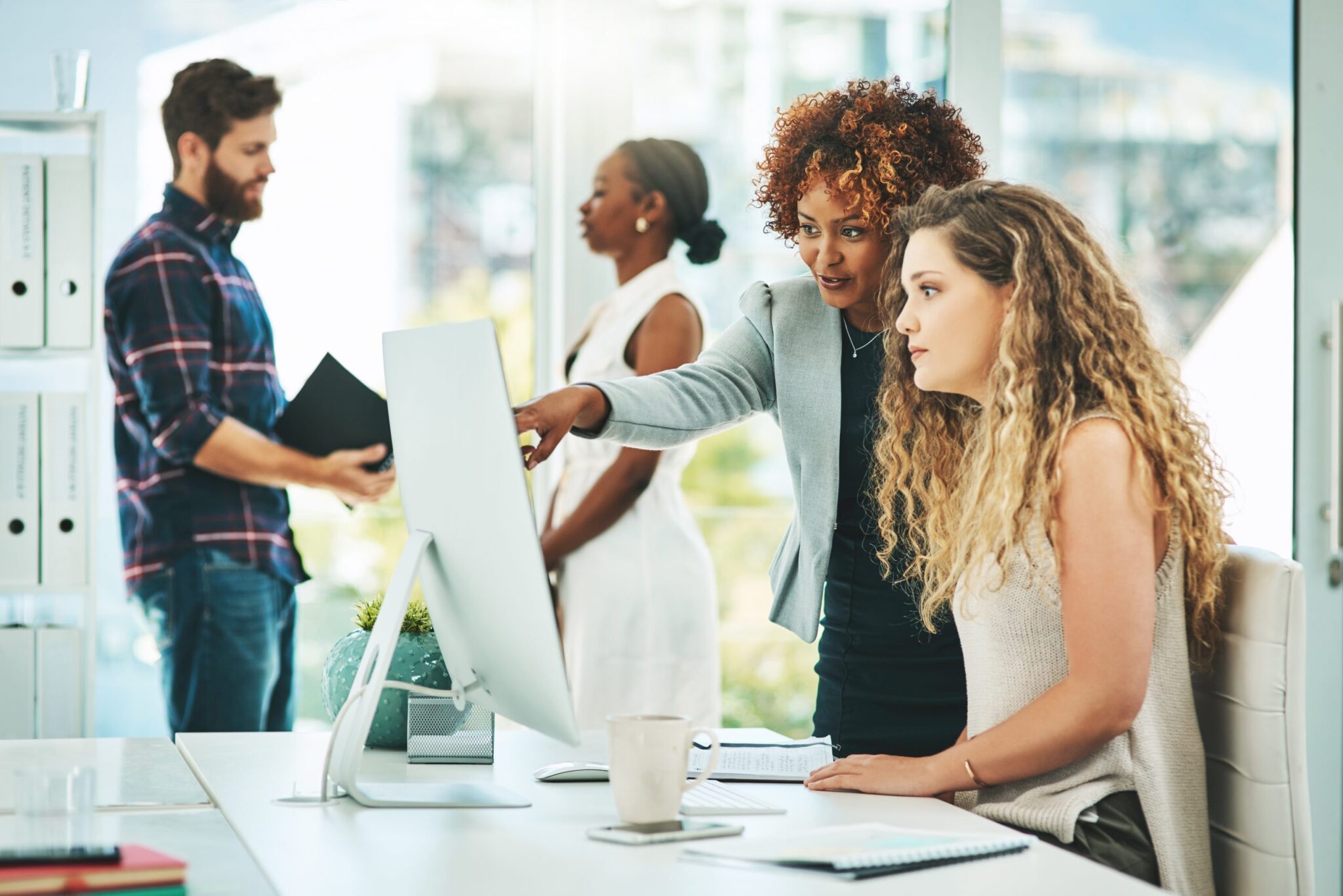 Shot of two businesswomen working together on a computer in an office with their colleagues in the background. Leading into 5 reasons to consider a post college internship