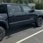 Side view of a black Toyota Tacoma SR5 pickup truck parked in a lot on a sunny day, showing the driver's side with black wheels and extended cab.