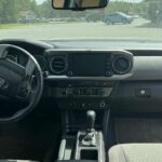 Front interior view of a Toyota Tacoma, showing the steering wheel, touchscreen display, climate controls, gear shift, and dashboard with Tacoma branding on the passenger side.