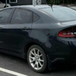 Rear-side view of a Black Dodge Dart parked on the side of a road, with trees and power lines in the background.