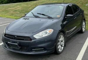 Front-side view of a Black Dodge Dart parked on the side of a road, with trees in the background.