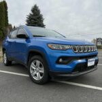 A bright blue Jeep Compass SUV parked in a paved parking lot on a cloudy day, with trees and a sign visible in the background.